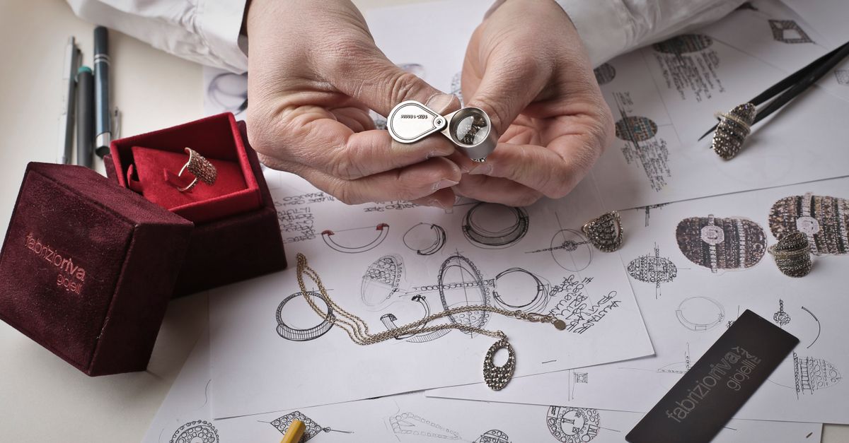 Close-up of hands inspecting a diamond ring with a loupe on a workbench filled with jewelry sketches.