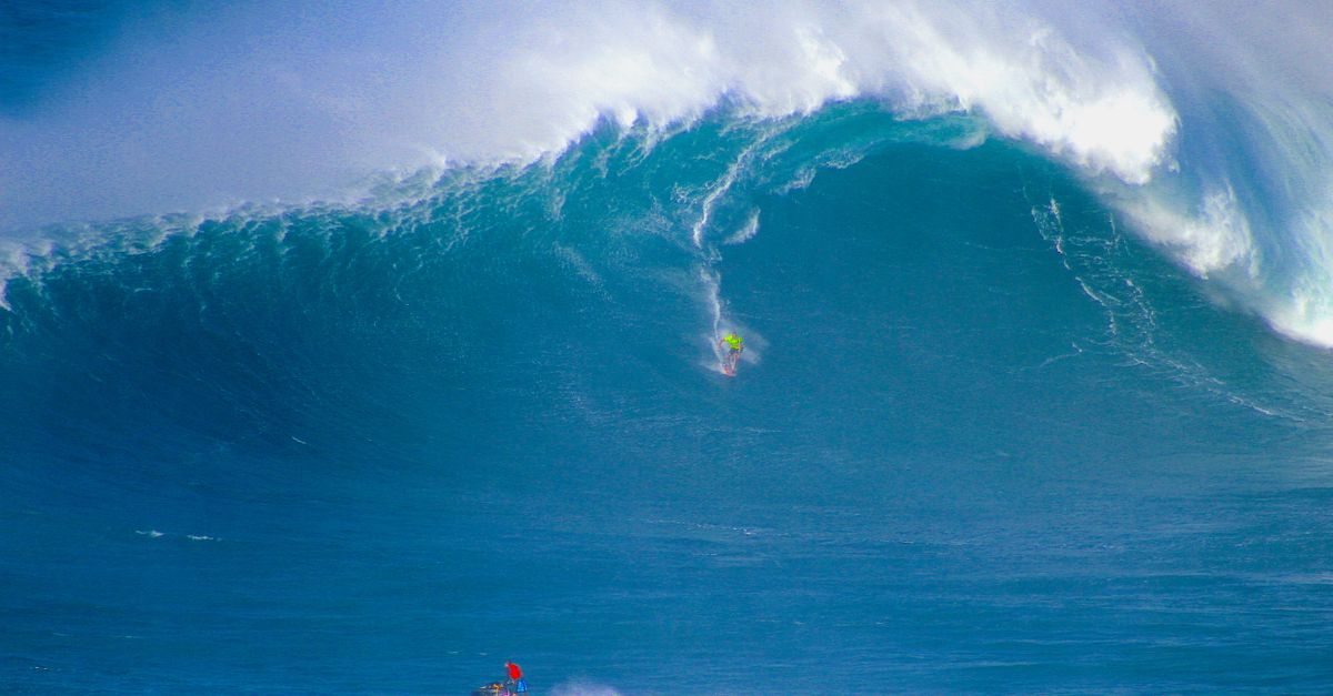 Thrilling scene of surfers tackling massive ocean waves with jet skis under a bright blue sky.