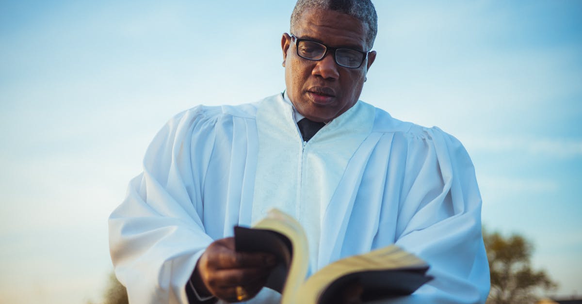 An adult pastor wearing glasses and a white robe reads a book outdoors under a blue sky.