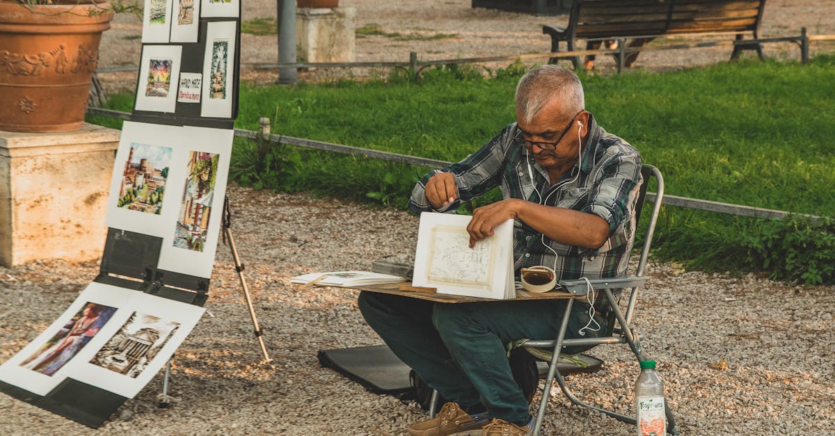 An elderly artist focused on sketching in a tranquil outdoor park setting, surrounded by his artwork.