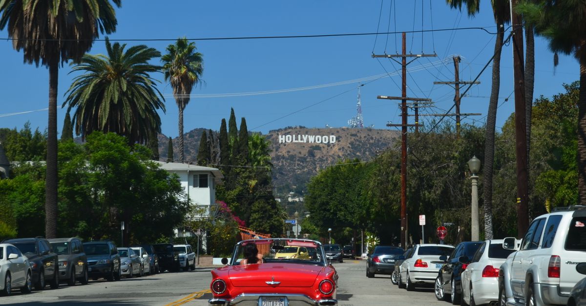 Red convertible driving down a sunny street towards the iconic Hollywood sign in Los Angeles.