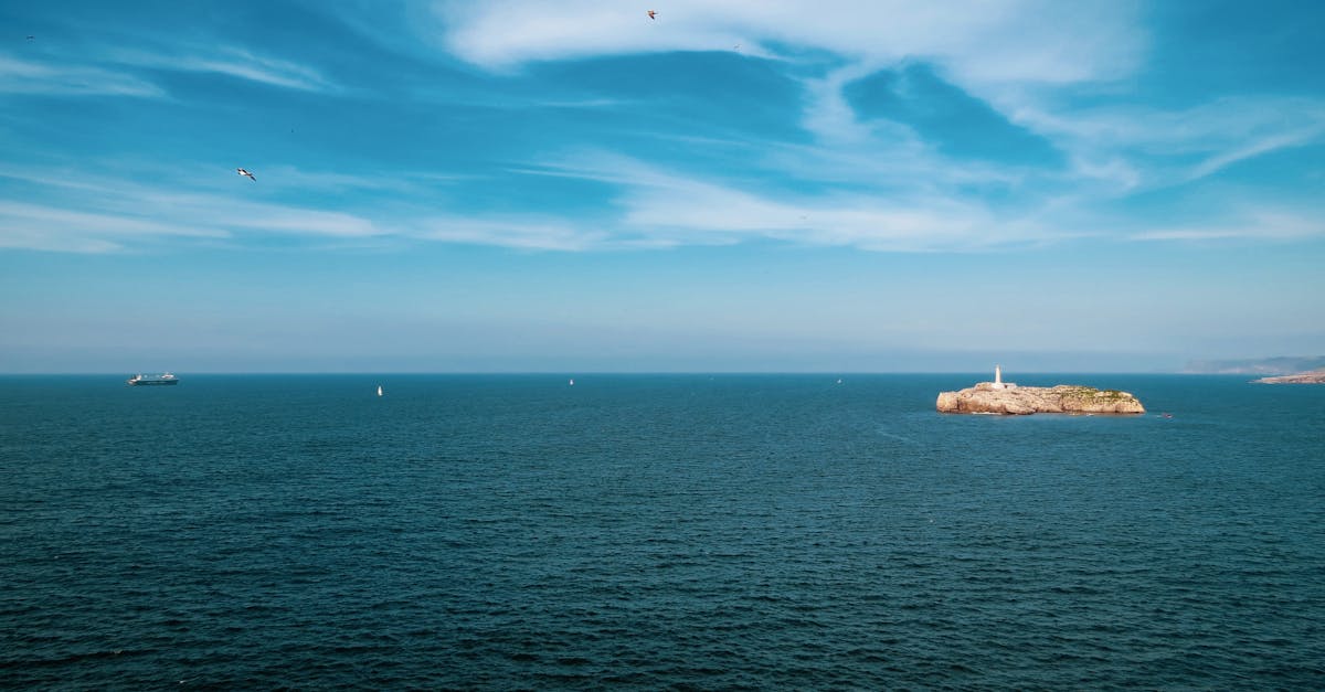 Wide-angle view of the ocean with a small island and lighthouse under a clear blue sky.