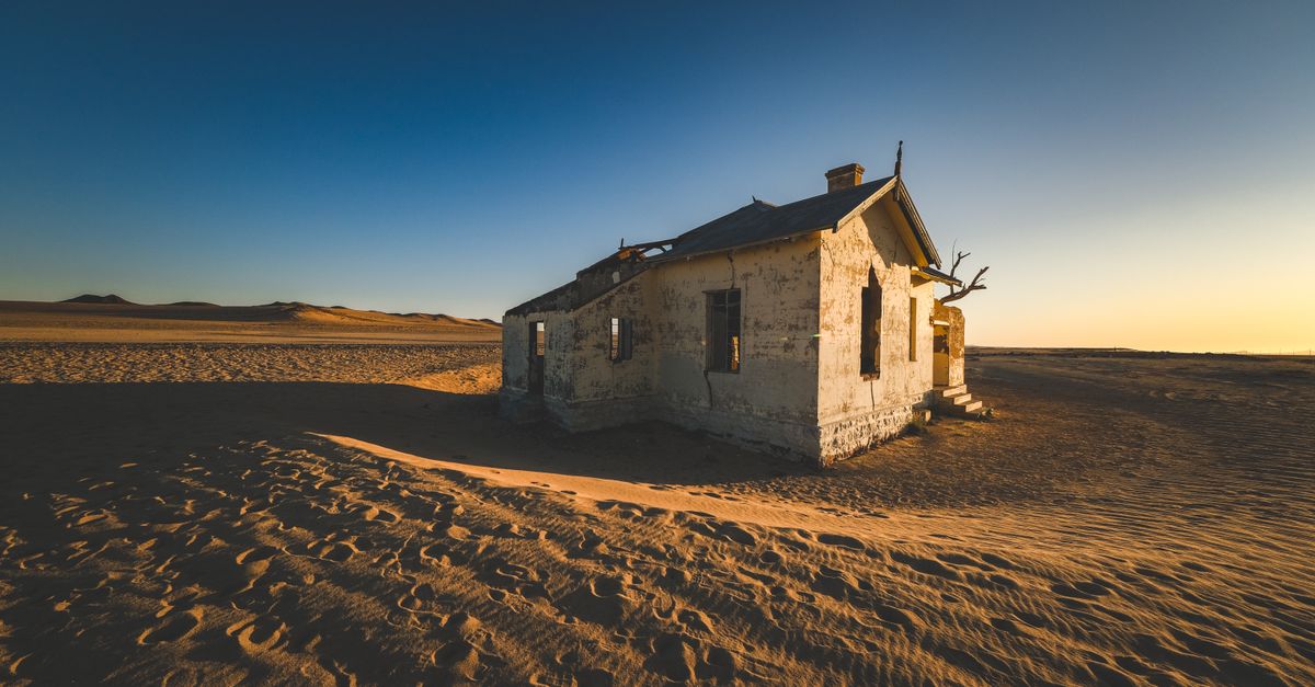 An abandoned house stands isolated in a desolate desert, surrounded by dunes at sunset.
