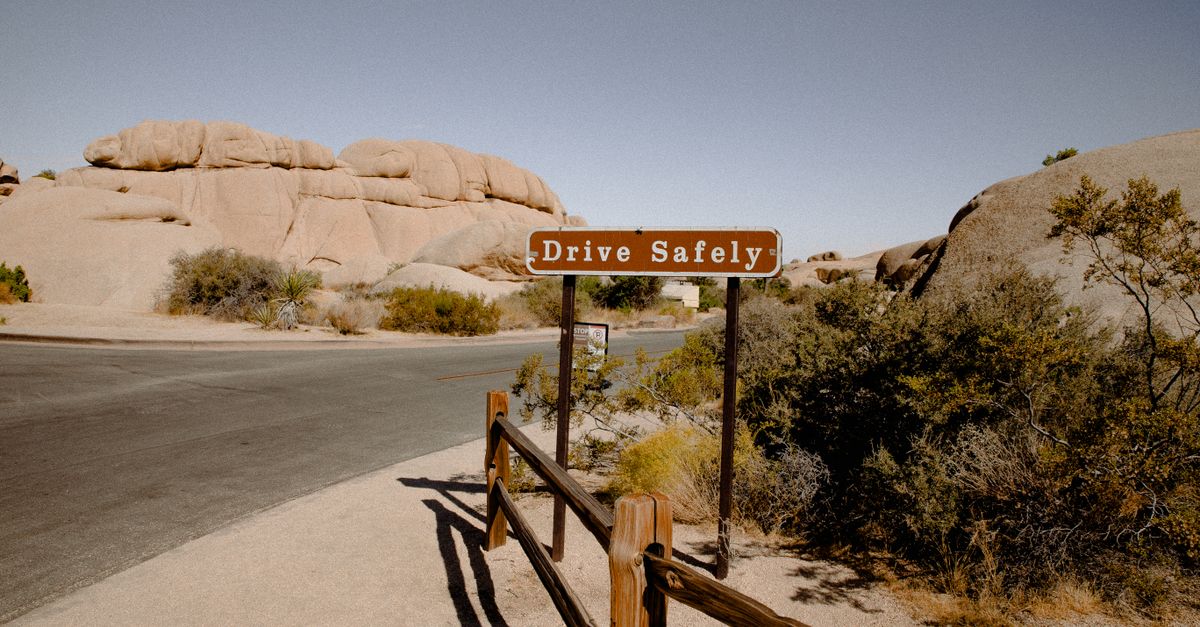 A scenic desert road with a 'Drive Safely' sign surrounded by rocky formations and dry vegetation.