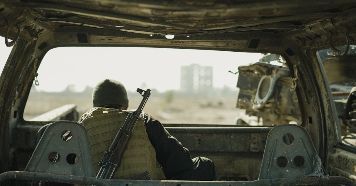 A soldier in a vest sits in an abandoned vehicle, observing a distant building.