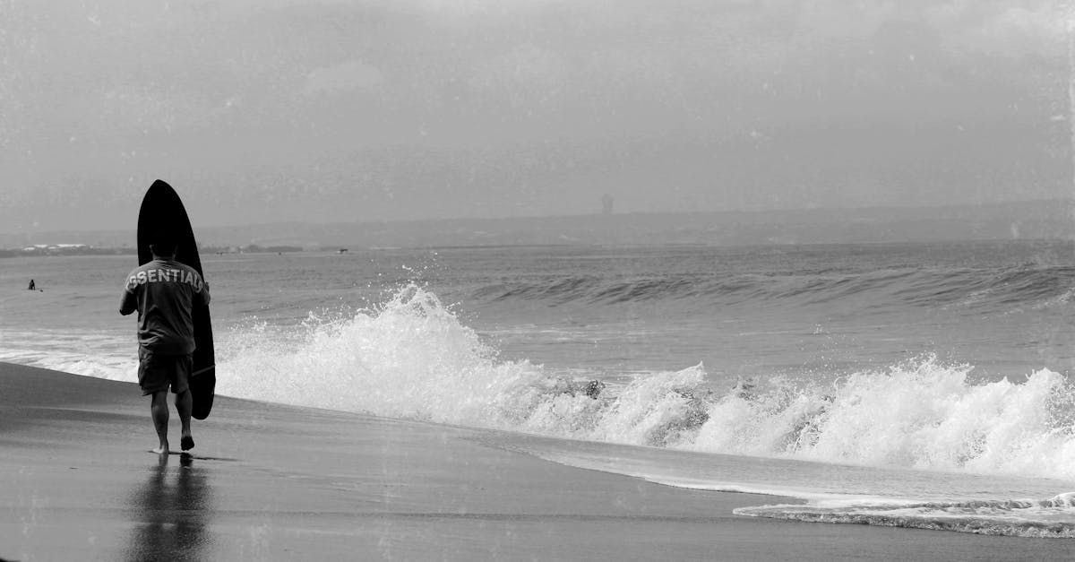Black and white image of a surfer walking on Bali beach with waves crashing.