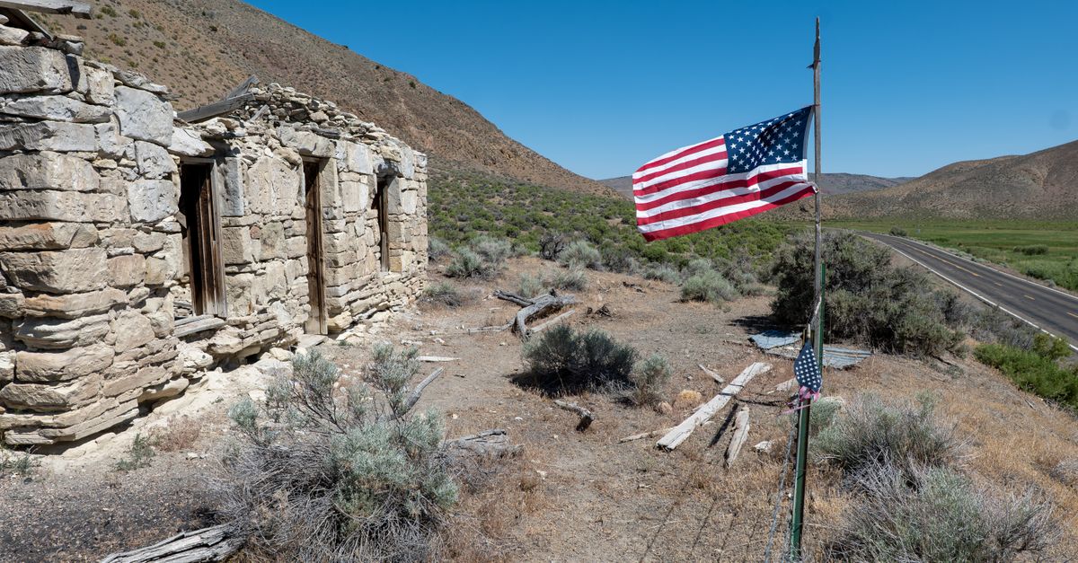 Rural landscape with an abandoned stone building and American flag under a clear blue sky.