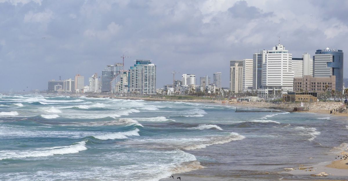 Waves crashing along the Tel Aviv shoreline with a view of modern skyscrapers under a cloudy sky.
