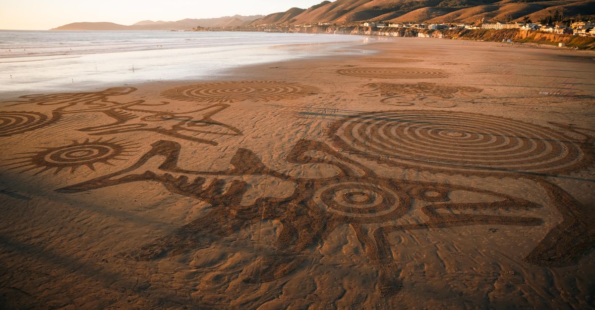 Aerial view of detailed sand art on a beach with scenic coastal hills at sunset.