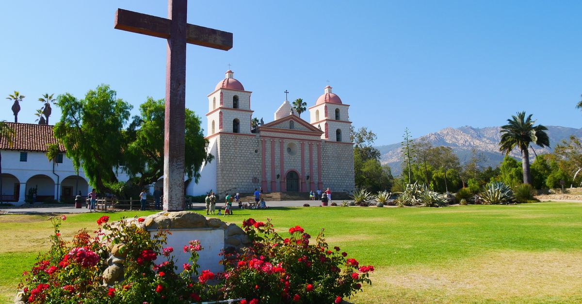 A sunny view of the Santa Barbara Mission with a cross and flowers in the foreground.