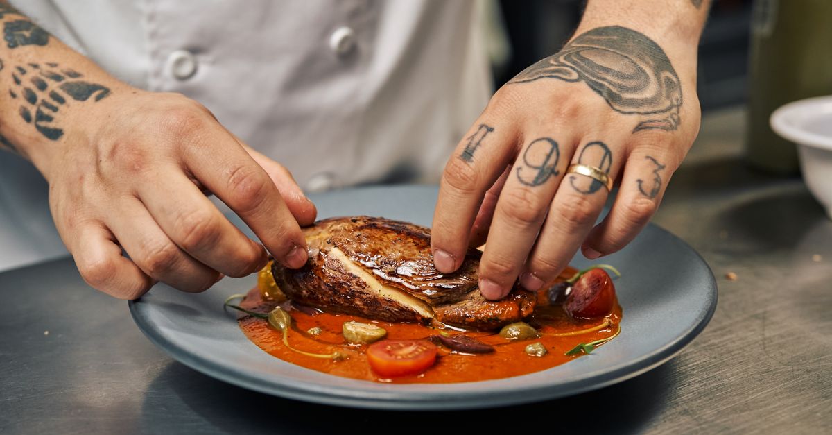 A chef with tattoos skillfully plates a gourmet dish in a professional kitchen.
