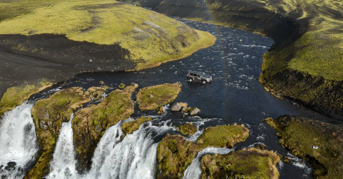 An off-road SUV navigates a river surrounded by lush Icelandic landscapes and waterfalls.