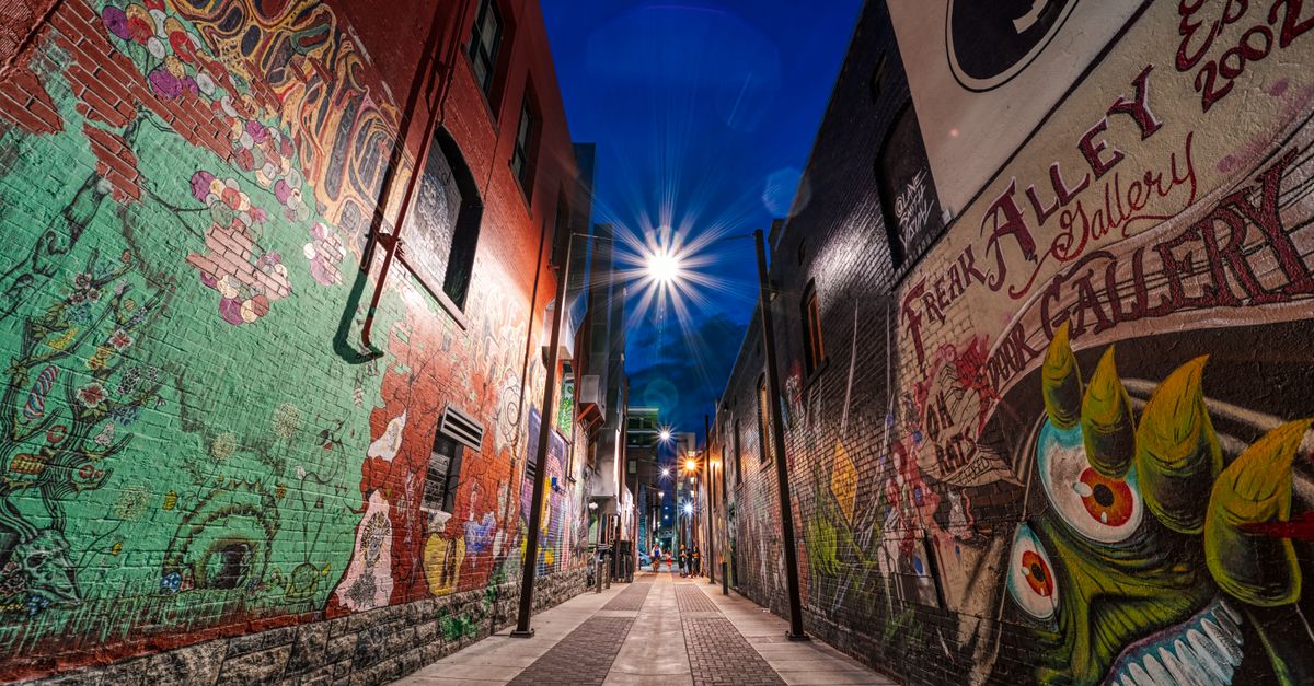 From below of empty walkway between aged houses with colorful graffiti walls under blue sky and shiny light at night