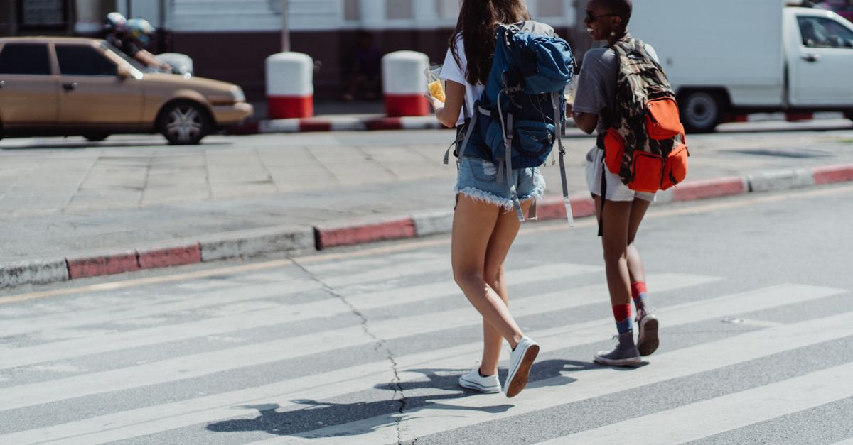 Two young women with backpacks crossing a city street in daylight, exploring urban life.