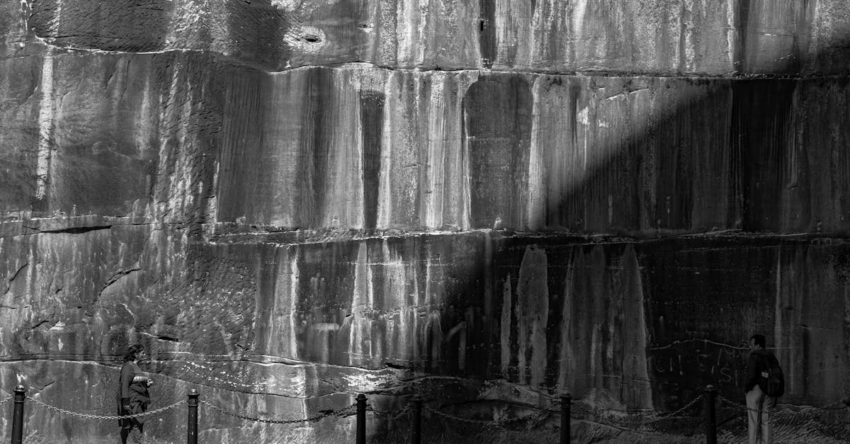 Black and white side view of faceless tourists walking on path near textured rocky cliff