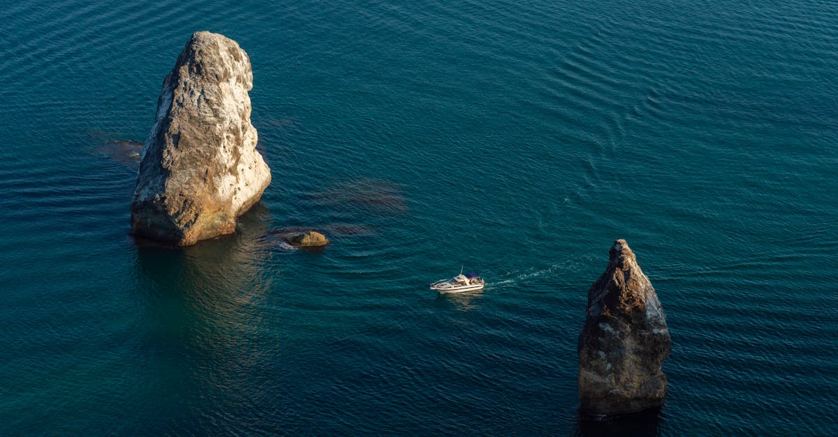 A yacht sails through striking rock formations on a calm blue sea, captured from above.