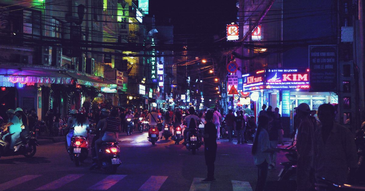 Pedestrians and motorcyclists on busy city street with bright illuminations at evening in Asian country