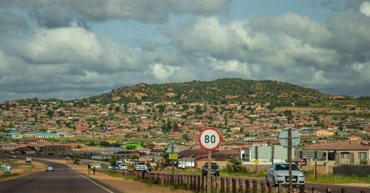 A vibrant countryside town nestled by mountains and a scenic road with vehicles under a blue sky.