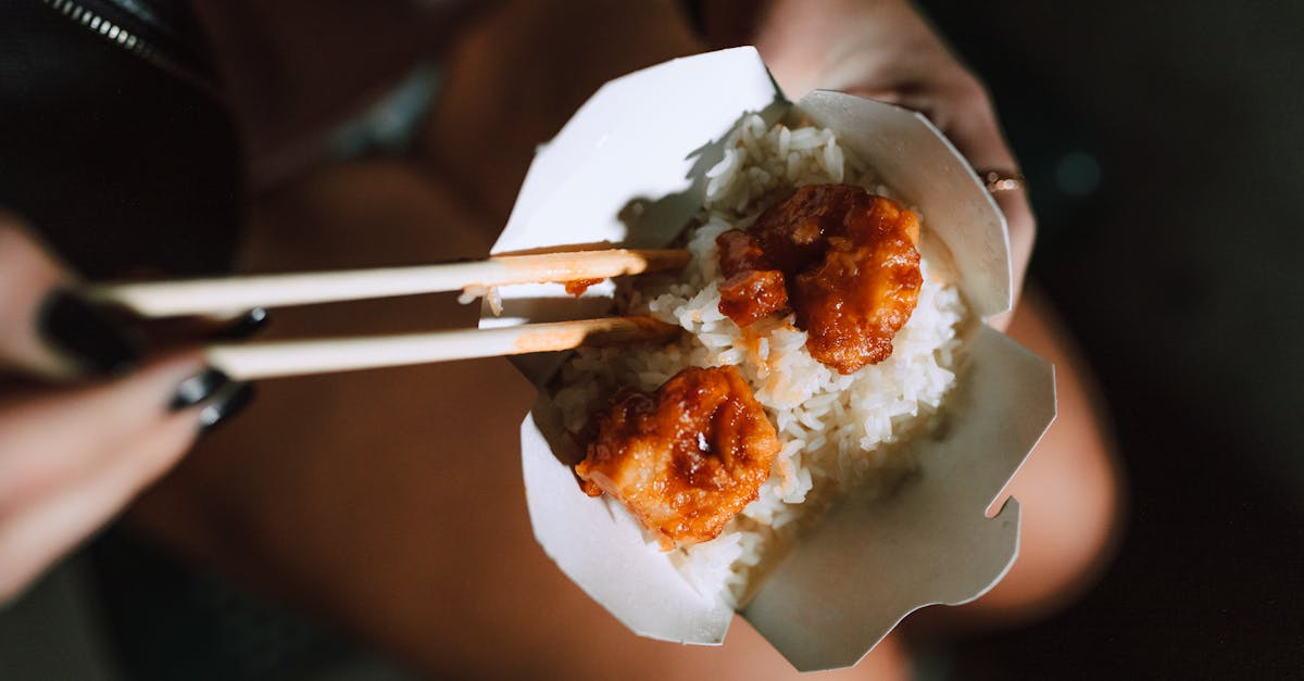 Fried shrimp with rice in a takeout box, held with chopsticks, exuding Asian culinary appeal.