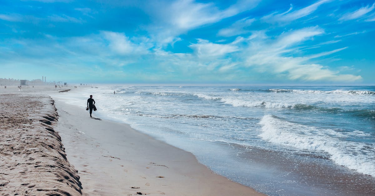 A surfer walking on a sandy beach with waves crashing under a blue sky, capturing coastal tranquility.